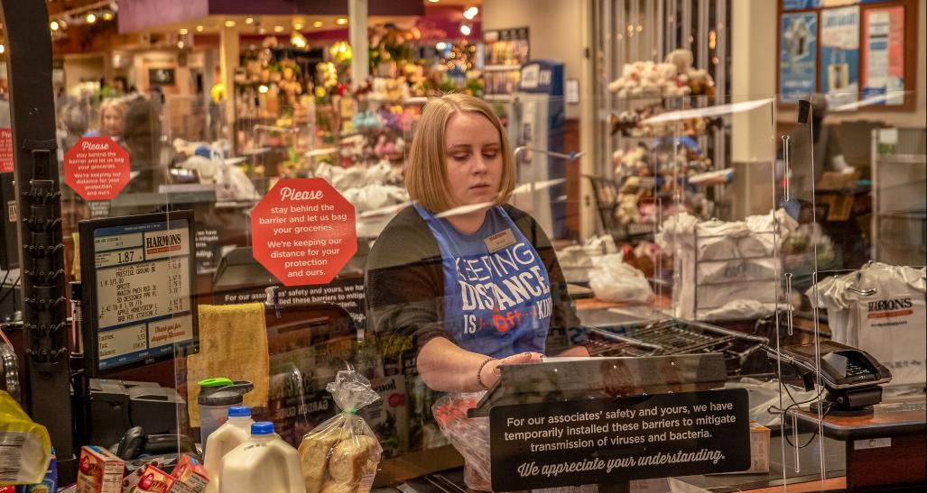 A grocery store clerk rings up items behind a plexiglass barrier